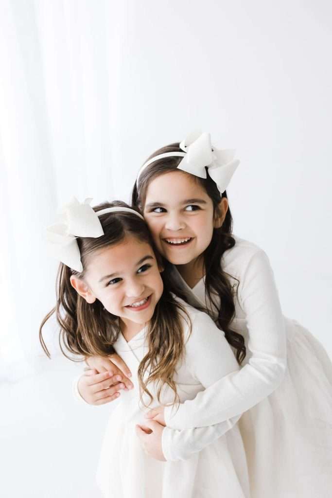 Two young girls in white dresses with white hair bows smile and hug each other against a plain white background.