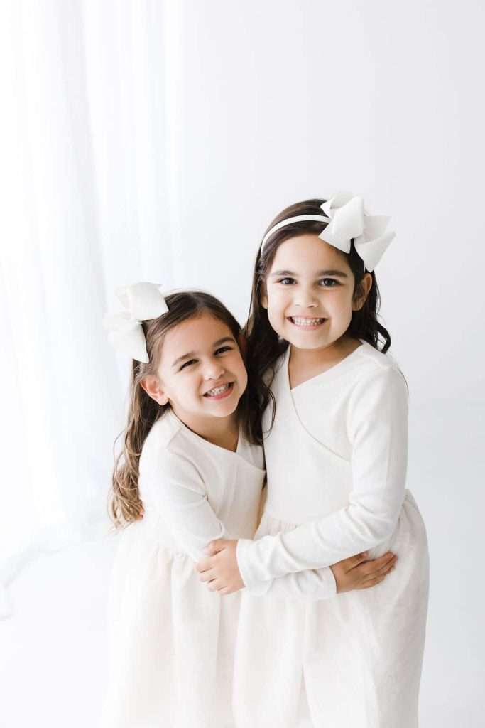 Two young girls with long brown hair, wearing matching white dresses and large white bows, stand close together and smile at the camera against a white background.