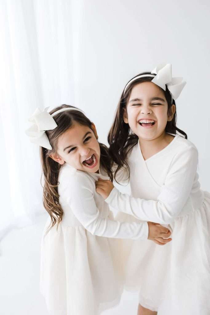 Two young girls in white dresses and matching headbands laugh together, standing closely in a bright, minimalist room.