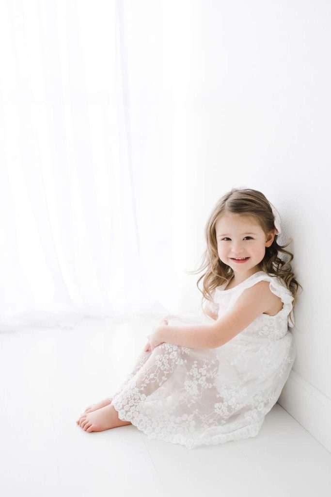 Young girl with long hair in a white lace dress sits barefoot on the floor against a white wall, smiling at the camera in a bright, softly lit room.