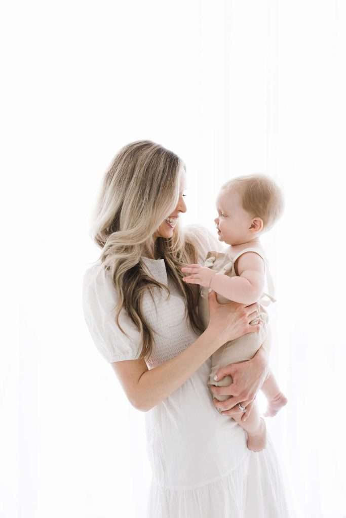 Woman in a white dress holds a baby and smiles at the child, who is looking back at her. The background is bright white.