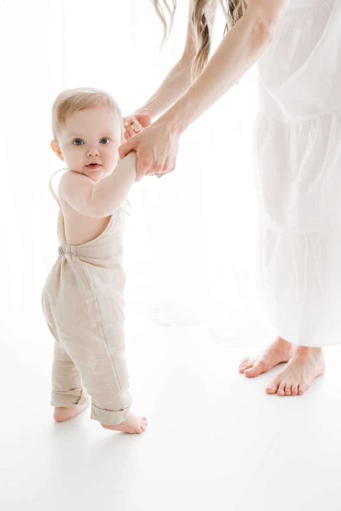 Baby in light overalls stands on a white floor while an adult in a white dress holds the baby’s hands to help them balance.