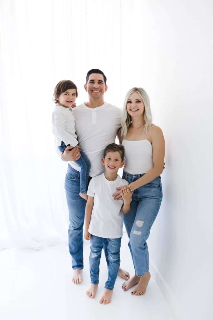 A family of four poses together in matching white tops and blue jeans against a white background.