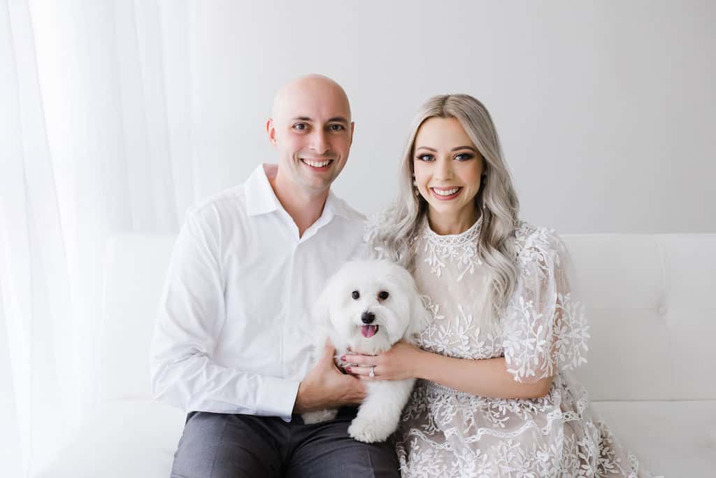 A man and a woman sit on a white couch, smiling at the camera, with a small white dog sitting between them.