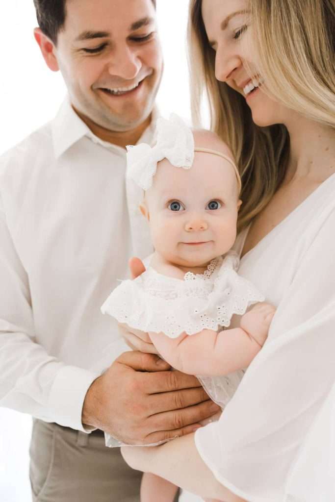 Smiling couple holds a baby dressed in white with a bow headband. All three are looking happy, and the background is bright and white.