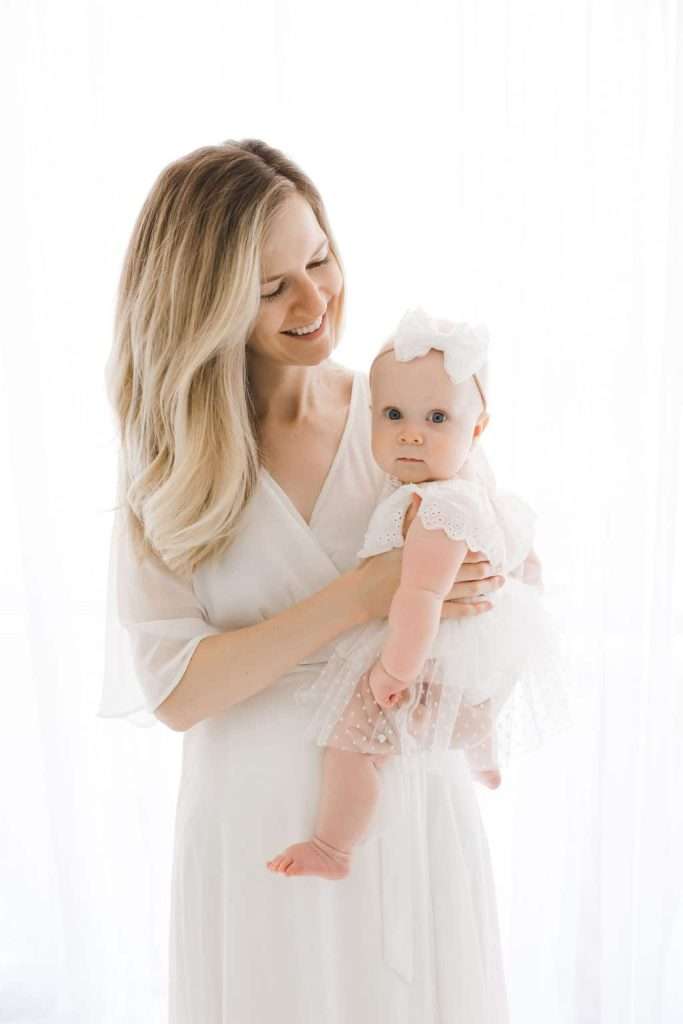 A woman in a white dress holds a baby dressed in white with a bow headband, both standing against a bright, white background.