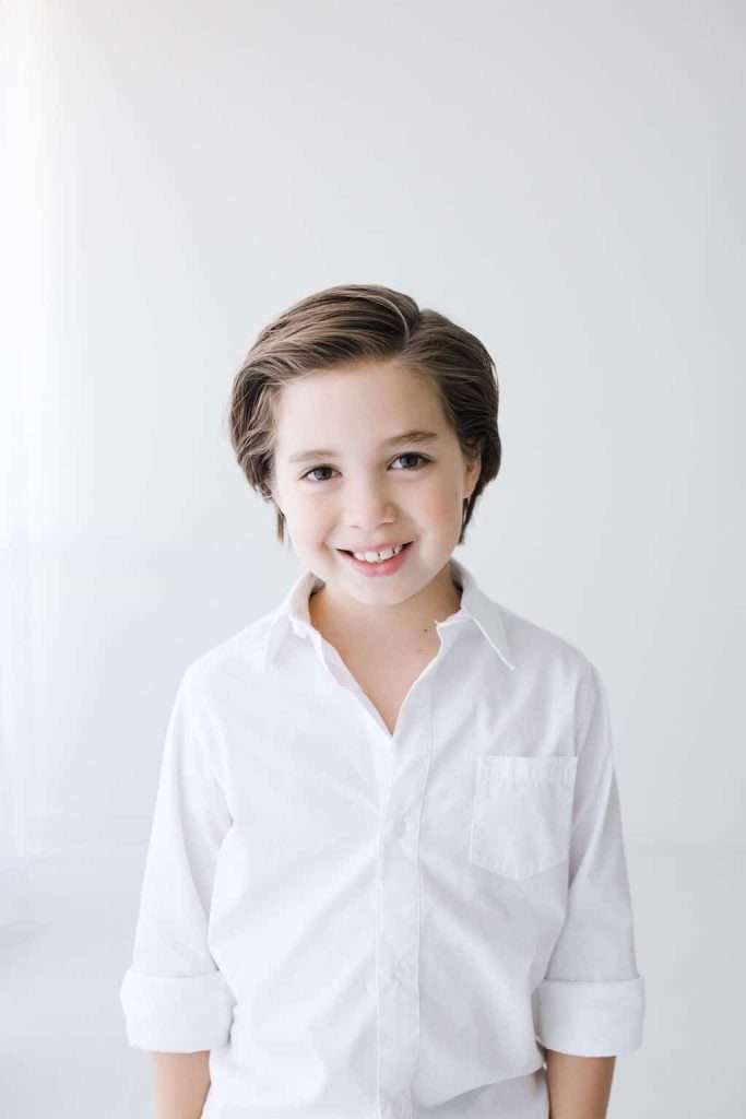 Smiling child with short brown hair wearing a white button-up shirt stands in front of a plain white background.