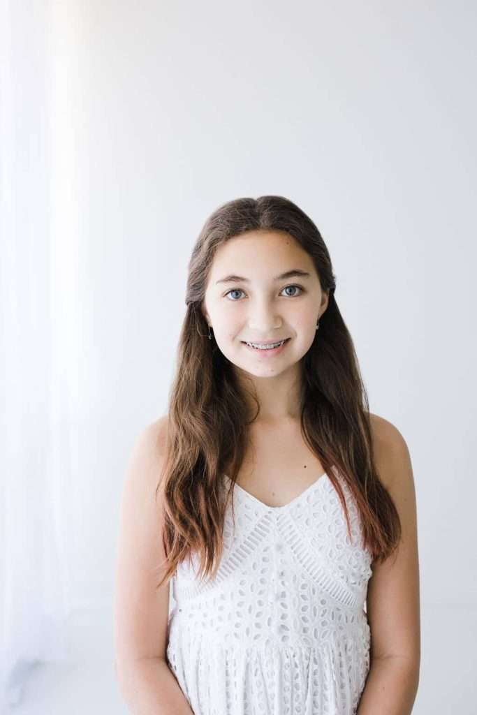 A young girl with long brown hair and braces stands indoors, wearing a sleeveless white dress, posed in front of a plain white background.