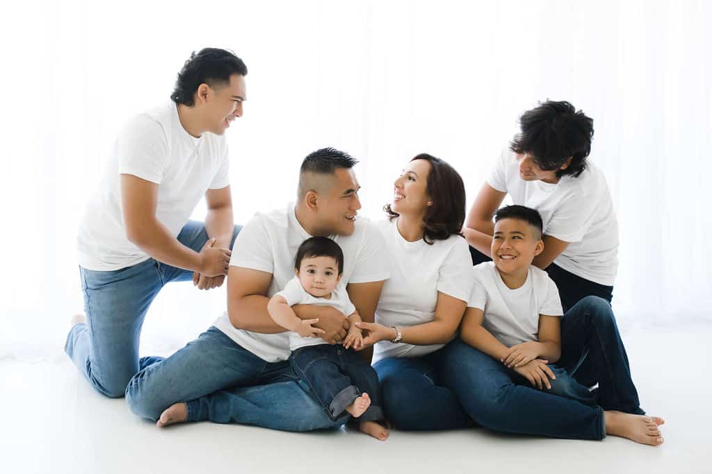 Six people, including two children, sit and kneel together on a white floor, all wearing white shirts and jeans, smiling and interacting with each other against a white background.