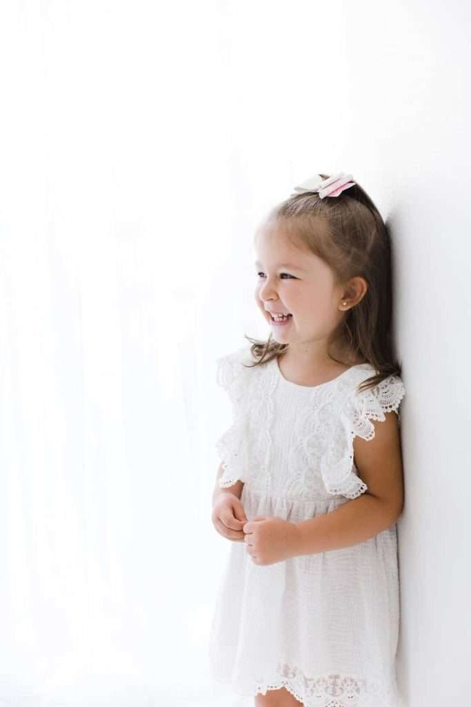 Young girl in a white lace dress stands against a white wall, smiling and looking to the side.