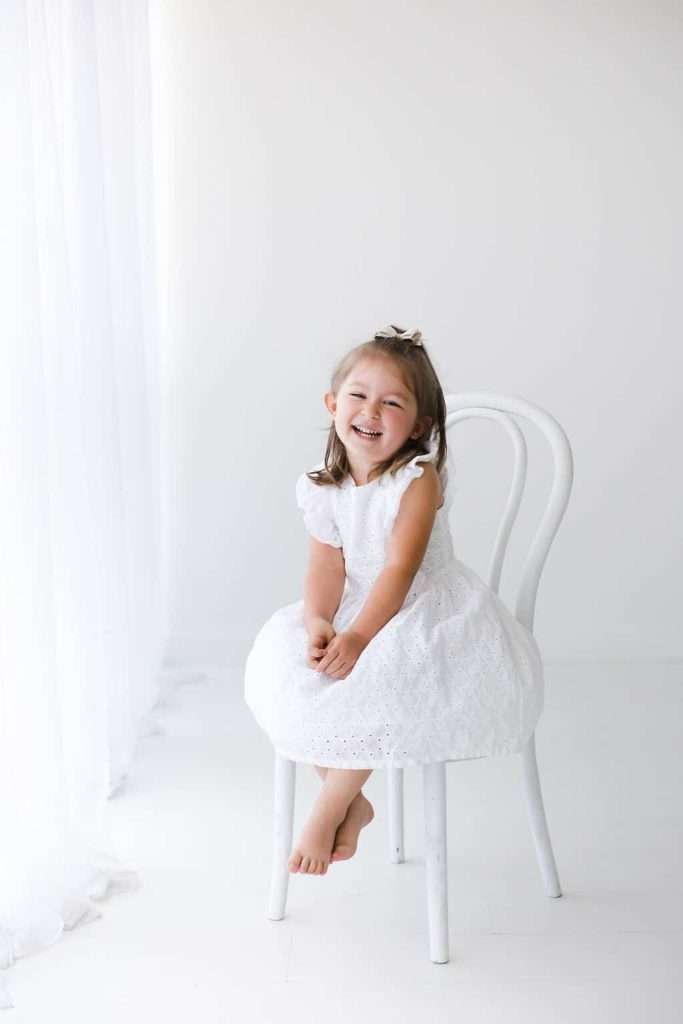 Young girl in a white dress sits barefoot on a white chair, smiling, with a white background and sheer curtains.