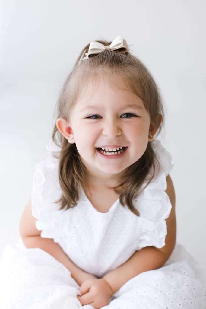 A young girl with light brown hair and a white bow smiles widely while wearing a white dress, sitting against a plain white background.
