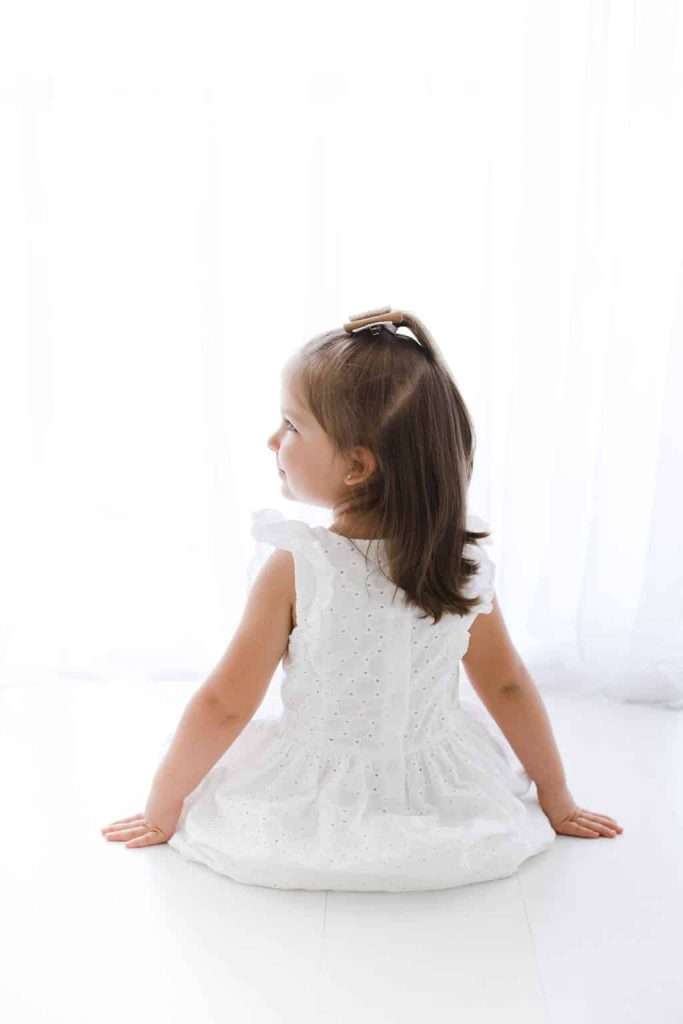 A young girl with brown hair sits on a white floor, facing away from the camera, wearing a white eyelet dress and a hair clip. Bright light comes from a window behind her.