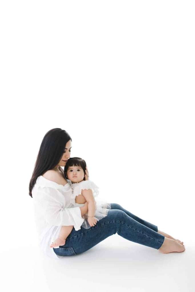 Woman sitting on floor in jeans and white top, holding a young child in a white dress on her lap, both looking forward against a plain white background.