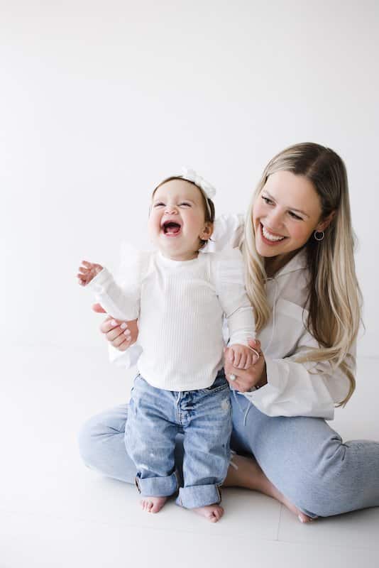 A woman sits on the floor holding a laughing toddler standing in front of her. Both are wearing white tops and blue jeans against a plain white background.