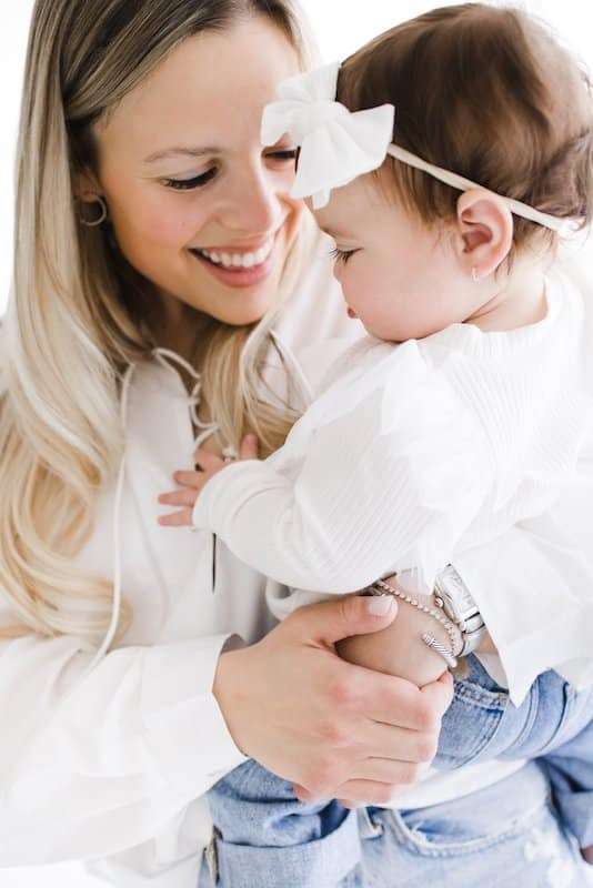 A woman with long blonde hair holds a baby wearing a white bow headband and white top. Both are smiling and dressed in light colors against a white background.
