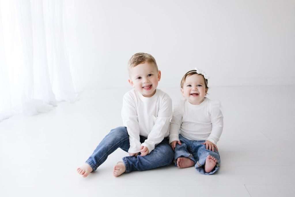 A young boy and a baby girl sit on the floor in matching white shirts and blue jeans, smiling at the camera in a bright, white room.