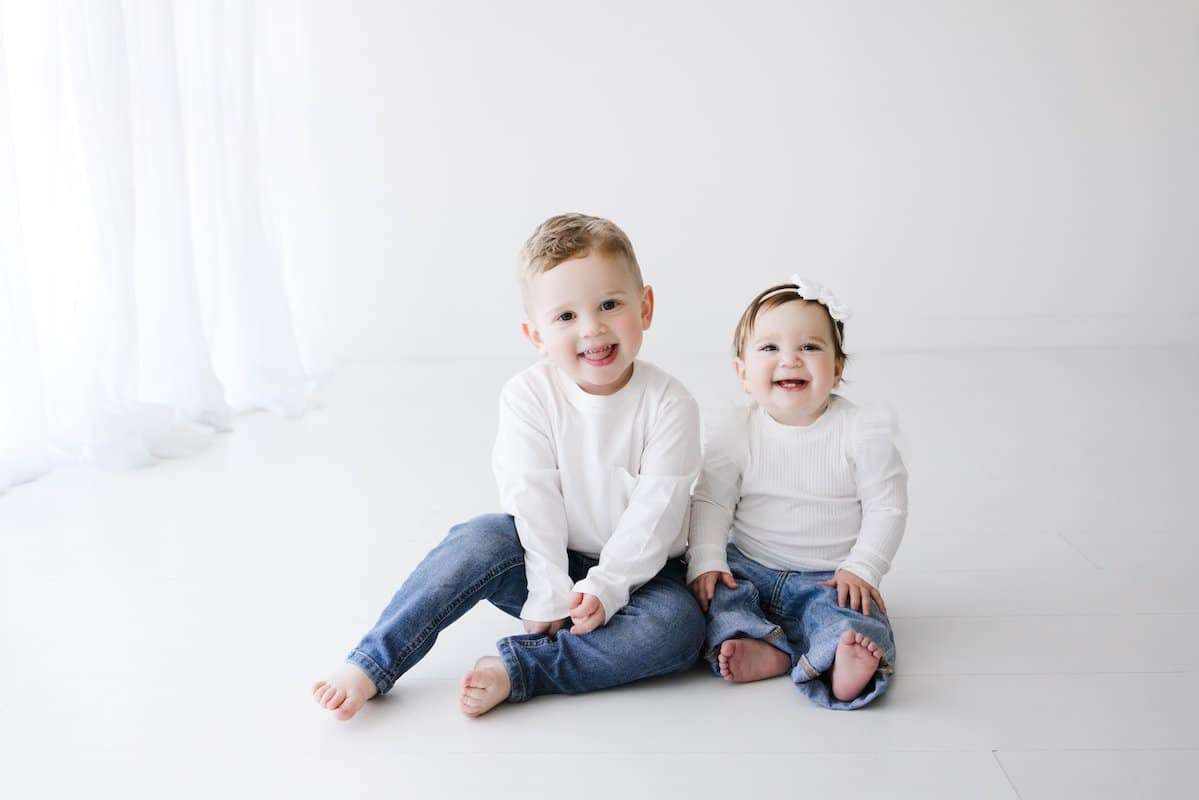 A young boy and a baby girl sit on the floor in matching white shirts and blue jeans, smiling at the camera in a bright, white room.