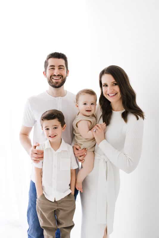 A family of four poses against a white background; two adults stand behind a young boy and a baby, all smiling and wearing light-colored clothing.