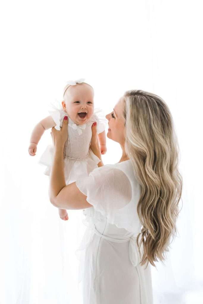 A woman with long blonde hair holds a smiling baby dressed in white, both facing each other against a bright white background.