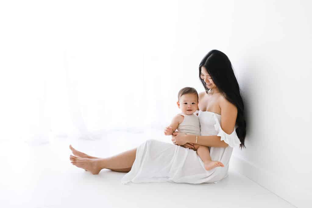 A woman in a white dress sits on the floor holding a baby in a light outfit. Both are barefoot against a white background.