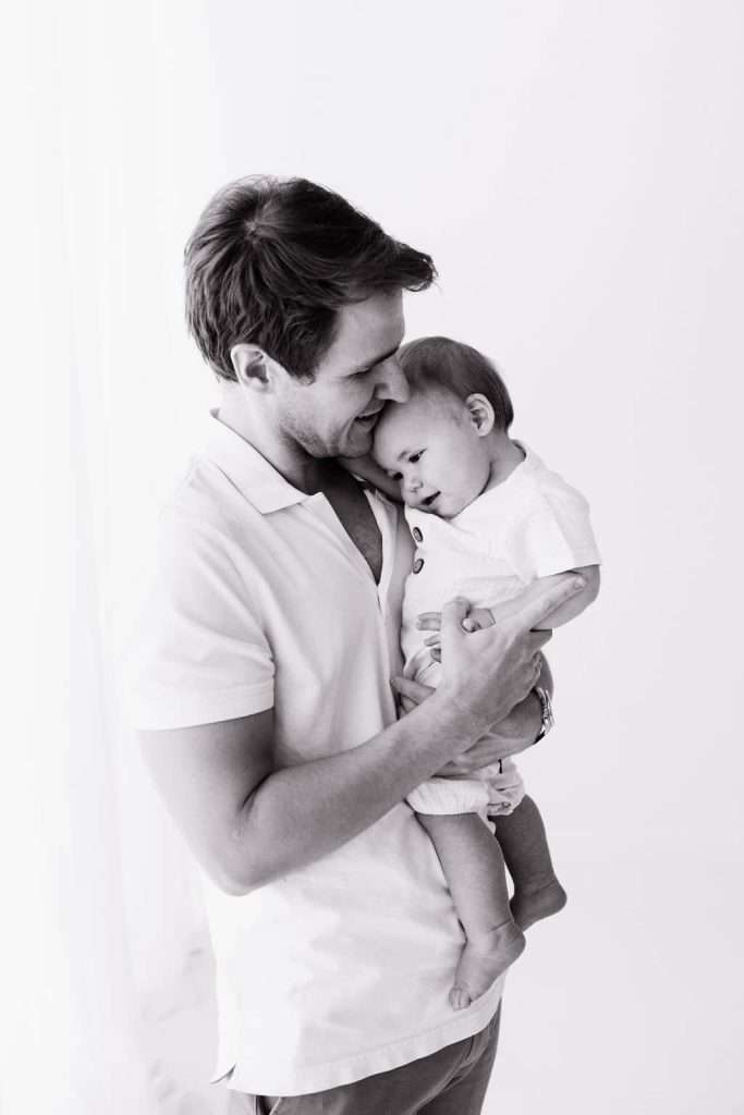 A man holding a smiling baby close to his chest, both dressed in light-colored clothes, on a plain white background.