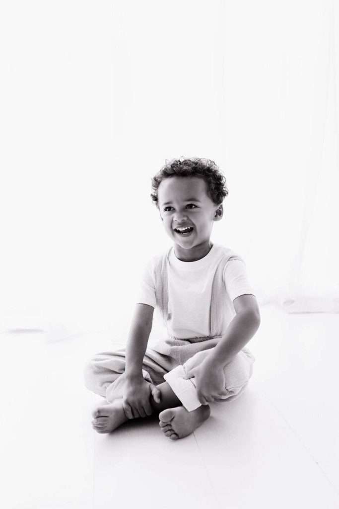 Black and white photo of a young child with curly hair, sitting barefoot on the floor and smiling, wearing light-colored clothes.