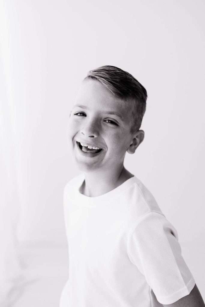 Black and white photo of a smiling boy with short hair, wearing a plain light-colored shirt, standing against a simple background.