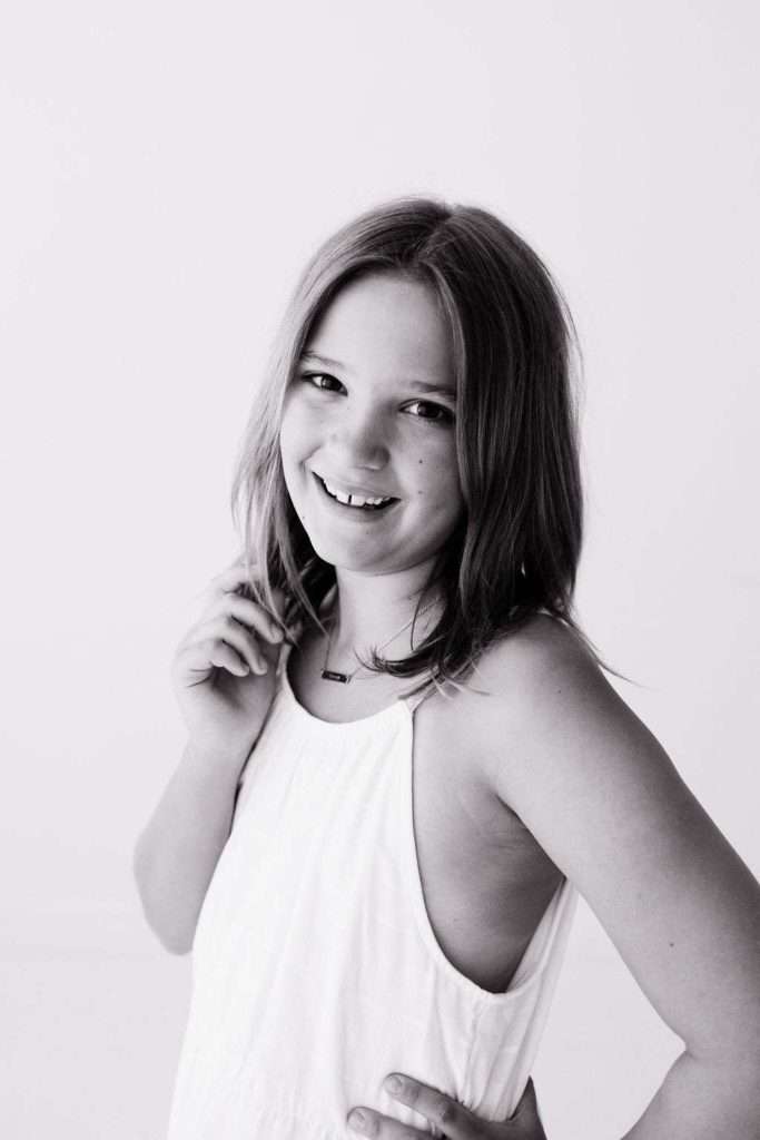 Black and white photo of a young girl with shoulder-length hair, wearing a sleeveless white top, smiling at the camera with one hand on her hip and the other near her neck.