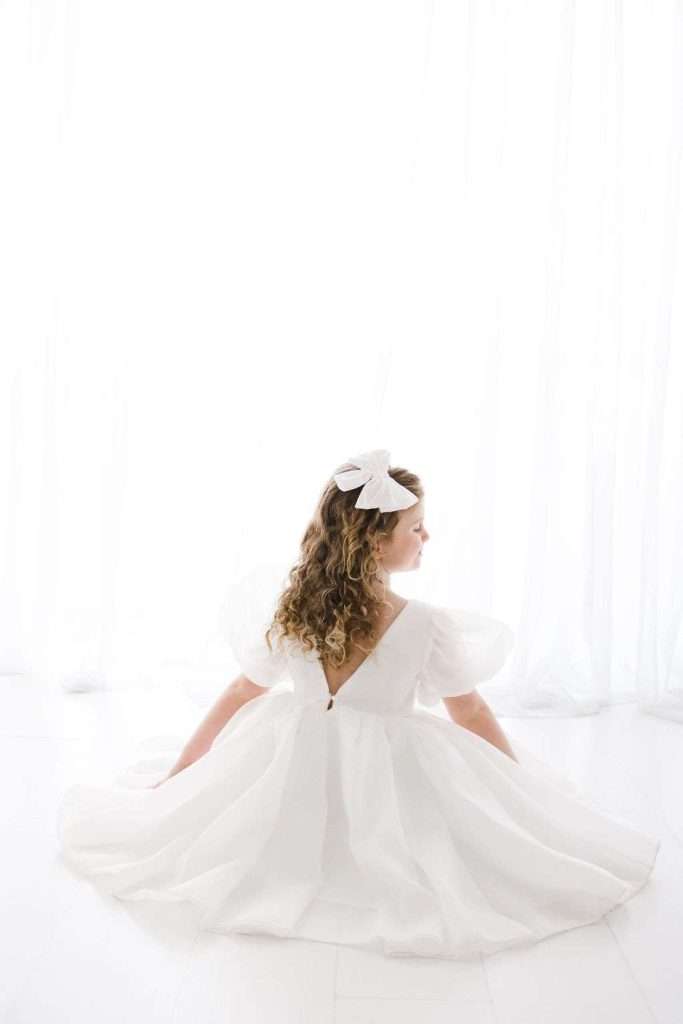 Girl with curly hair sits on the floor in a white dress and large white hair bow, facing away from the camera toward sheer white curtains.