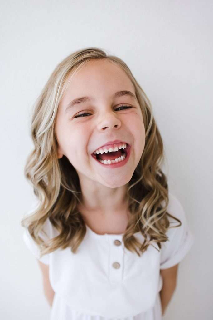 Young girl with wavy blonde hair wearing a white top is smiling widely and looking at the camera against a plain white background.
