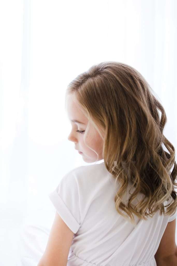 Young girl with wavy brown hair wearing a white shirt sits turned to the side, gazing downward, with a soft white background.