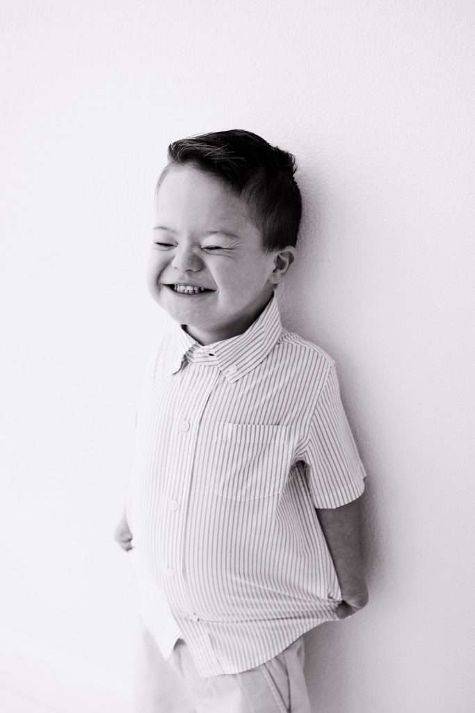 Young boy with closed eyes and a big smile stands against a white wall, wearing a light-colored, striped button-up shirt.