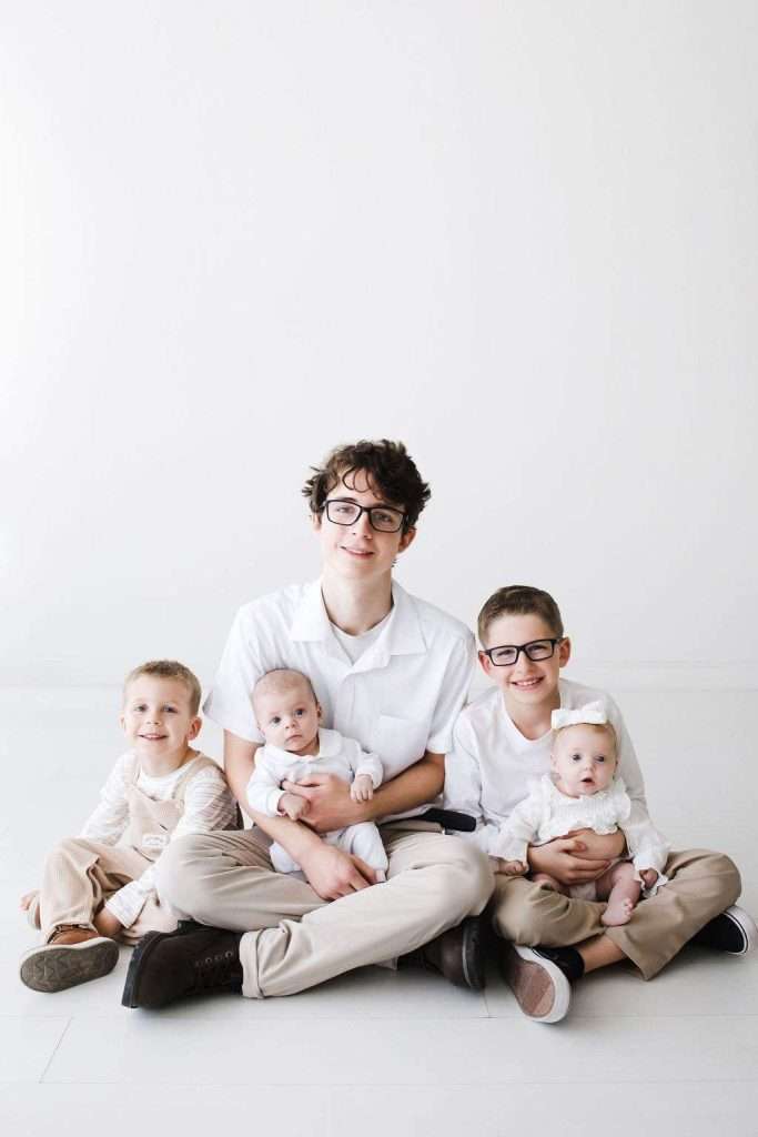 Five children in light-colored outfits sit on the floor against a plain white background, with the oldest child in the center holding two infants.