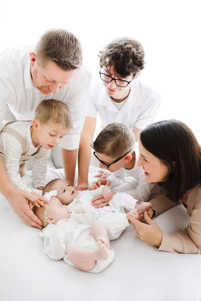 A family of six, including two adults, three children, and a baby, all dressed in white, gather closely on a white surface, smiling and interacting with the baby.