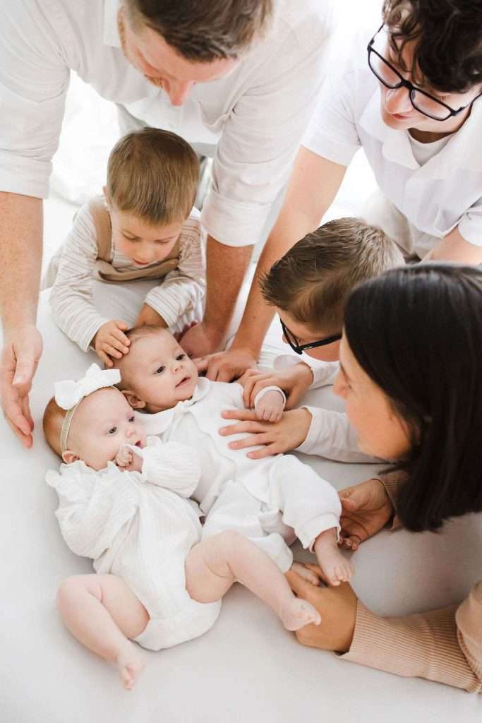 Two babies lie on a white surface, surrounded by four adults and one child, all looking at and interacting with the babies.
