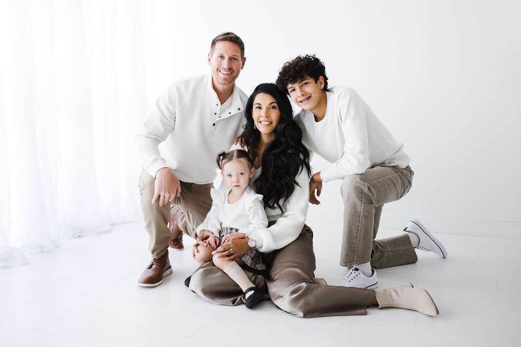Four people pose together in a brightly lit, white room. Two adults and two children are dressed in coordinating neutral outfits, sitting and kneeling on the floor, all facing the camera.