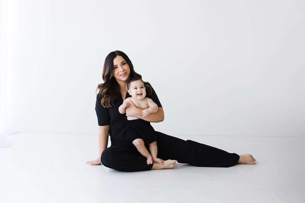 Woman in black outfit sits on the floor with a smiling baby on her lap against a plain white background.