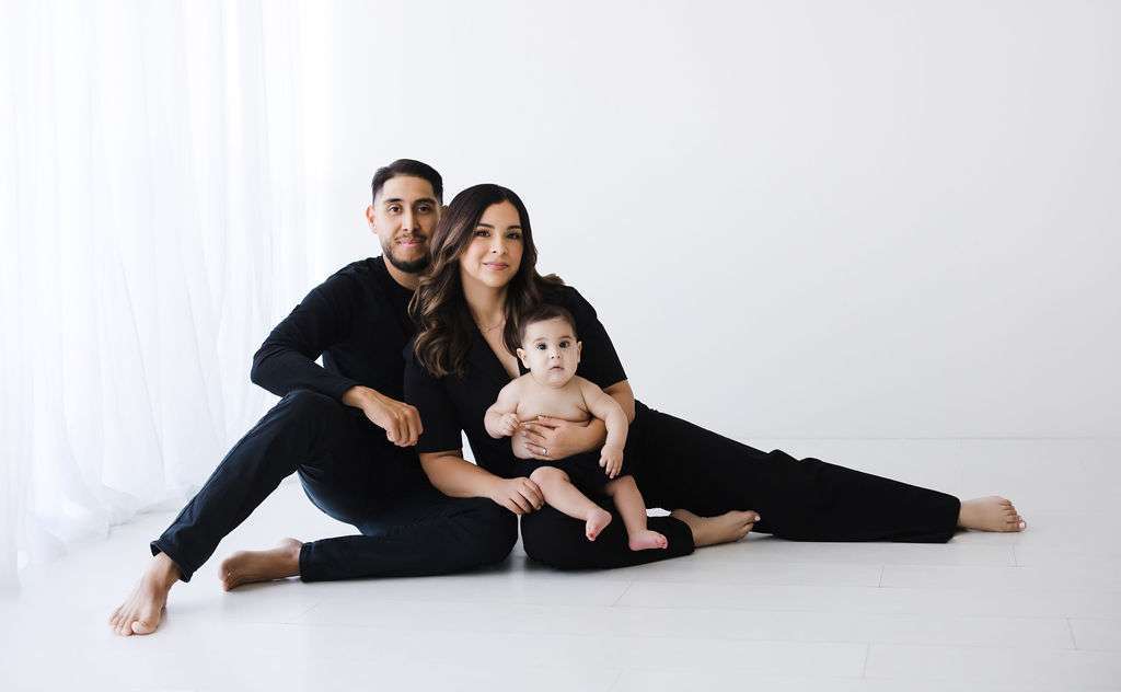 A man and woman dressed in black sit on the floor with a baby sitting on the woman’s lap, against a plain white background.