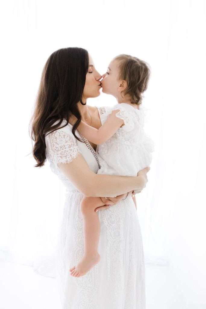 Woman in a white lace dress holds and kisses a young child, also in a white dress, against a bright white background.