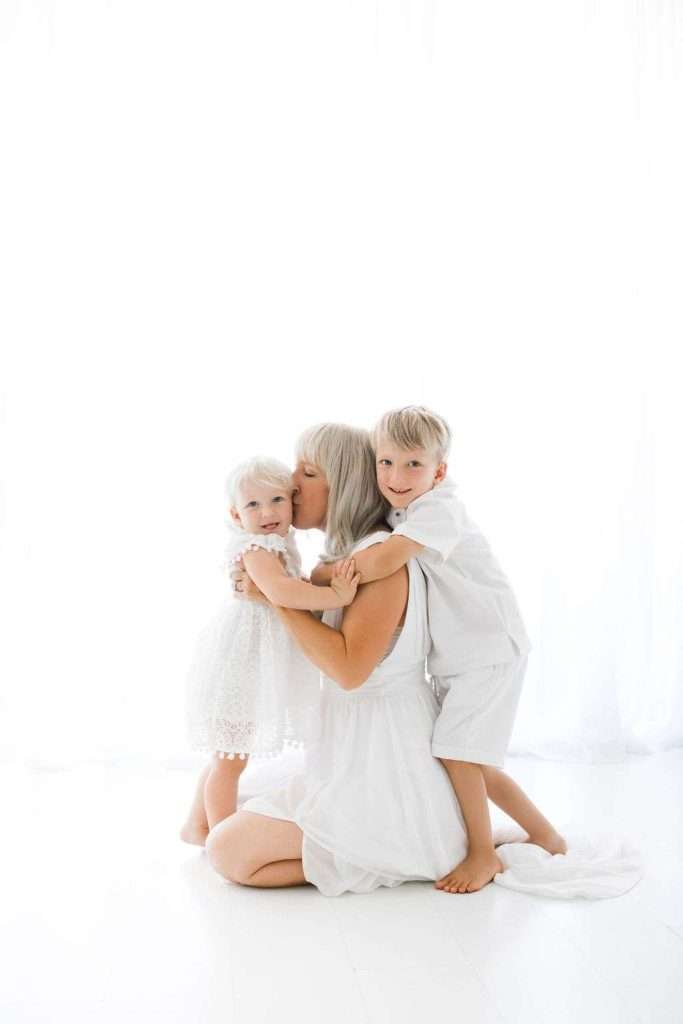 A woman in a white dress kneels on the floor, hugging a young boy and kissing a toddler girl. All are dressed in white, with a bright white background.