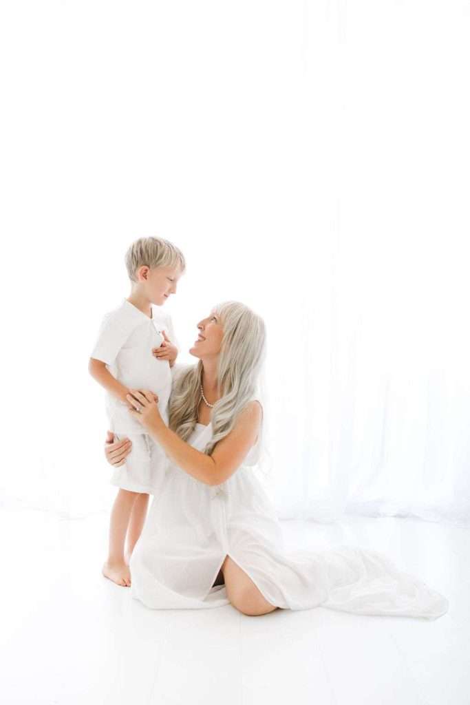 A woman with long gray hair kneels in front of a young boy. Both are dressed in white and smiling at each other in a bright, white room.