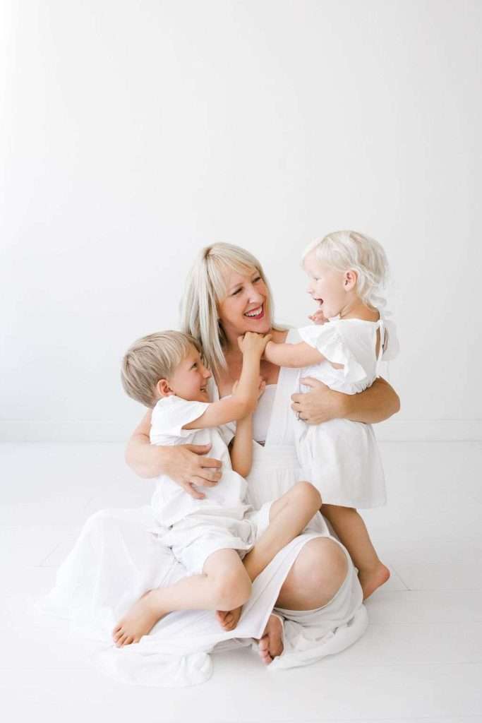 A woman in a white dress sits on the floor, smiling and embracing two young children dressed in white against a plain white background.