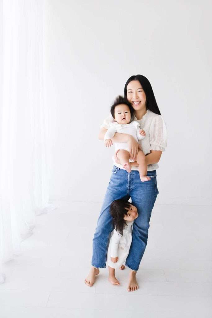 Woman stands barefoot holding a baby, with a toddler hugging her leg. All are dressed in light-colored clothes in a bright, minimal room.