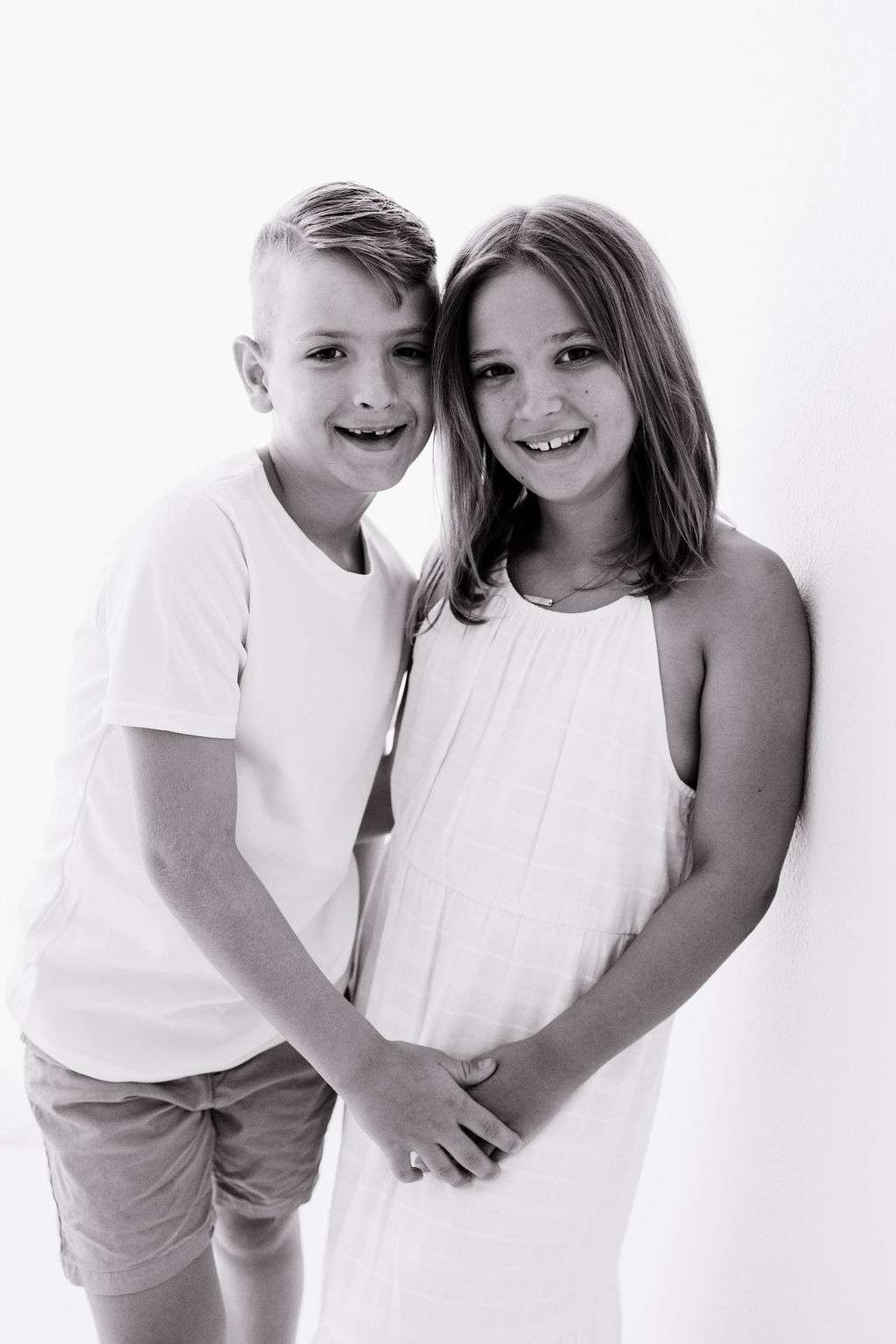 Two children standing close together, smiling at the camera. The boy wears a t-shirt and shorts, and the girl wears a sleeveless dress. The image is in black and white.