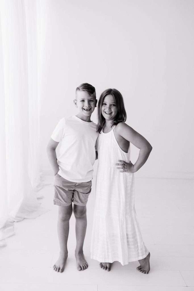 Two children stand together barefoot in a bright room, one wearing a t-shirt and shorts, the other in a sleeveless dress. Both are smiling at the camera.