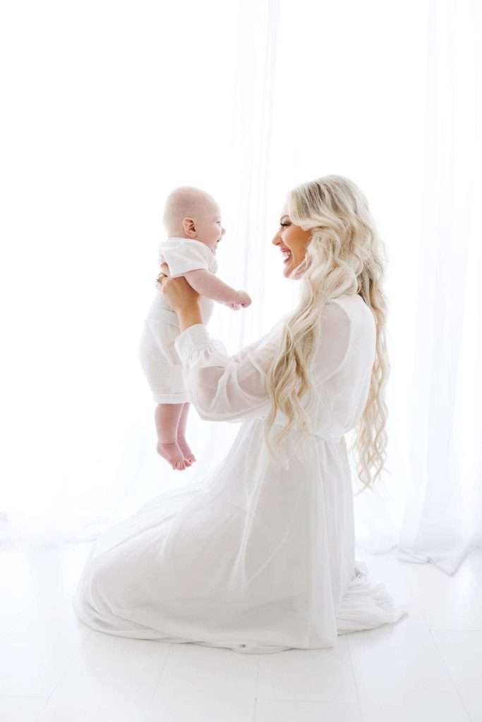 A woman in a white dress kneels on the floor, holding a smiling baby in white clothes up in front of her against a bright white background.