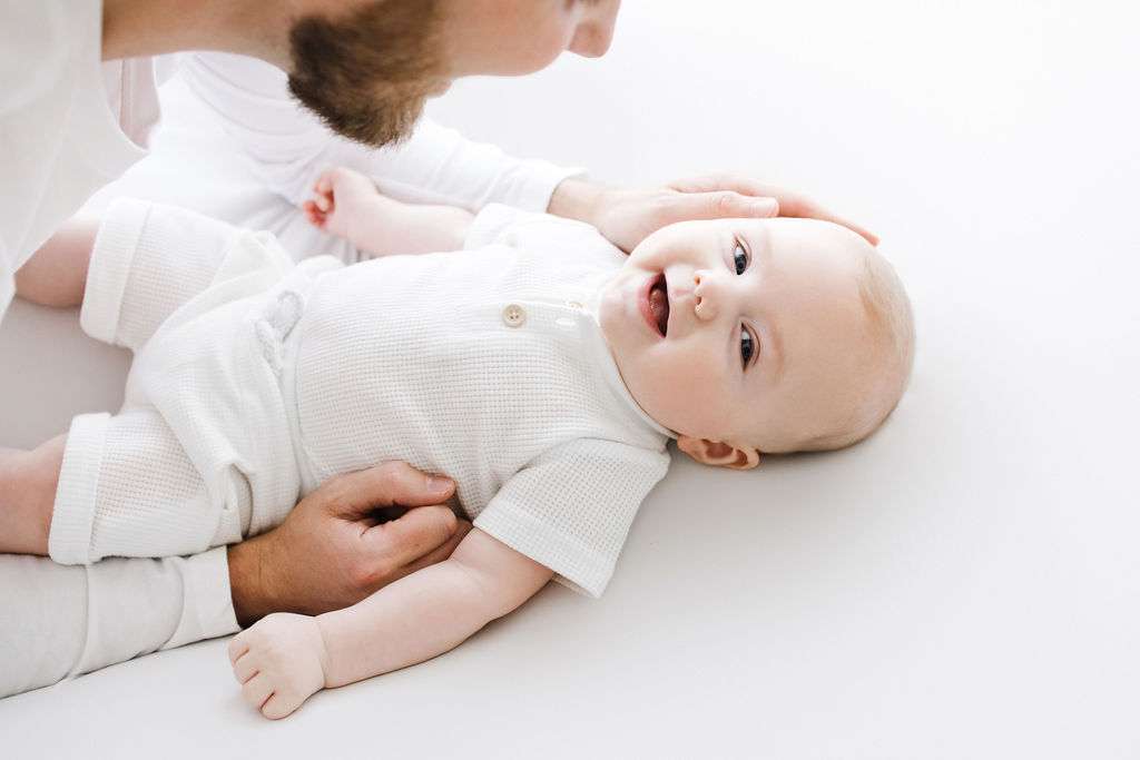 An adult gently cradles the head of a smiling baby lying on a white surface. Both are dressed in light-colored clothing and are facing each other.