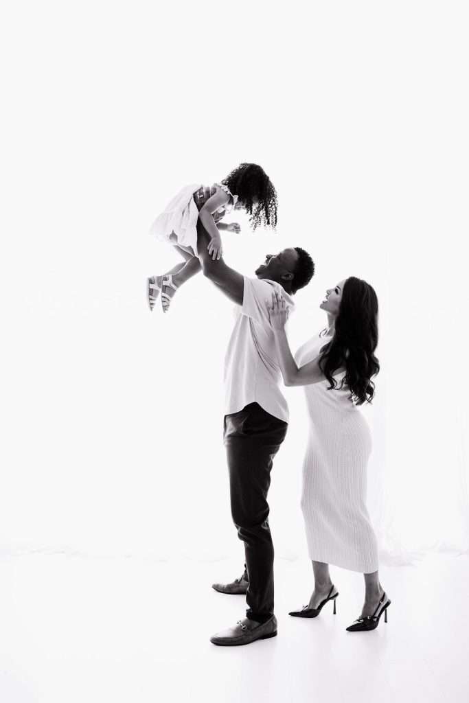 A man lifts a young girl in the air while a woman stands beside them, smiling. All three are dressed in light clothing against a white background.