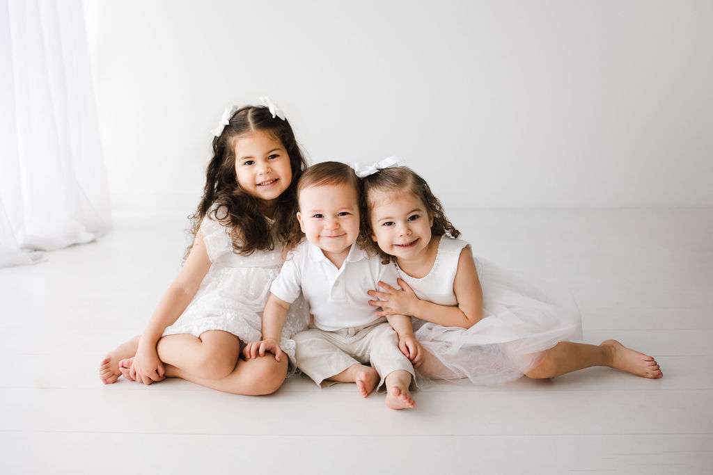 Three young children wearing white clothes sit on the floor, smiling at the camera against a plain white background.